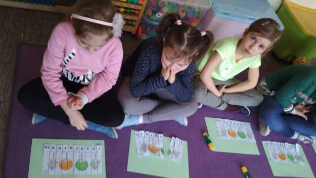 Kodowanie na dywanie… czyli początki programowania Dzieci siedzące po turecku na dywanie rozwiązują zadanie - układają pocięte paski papieru