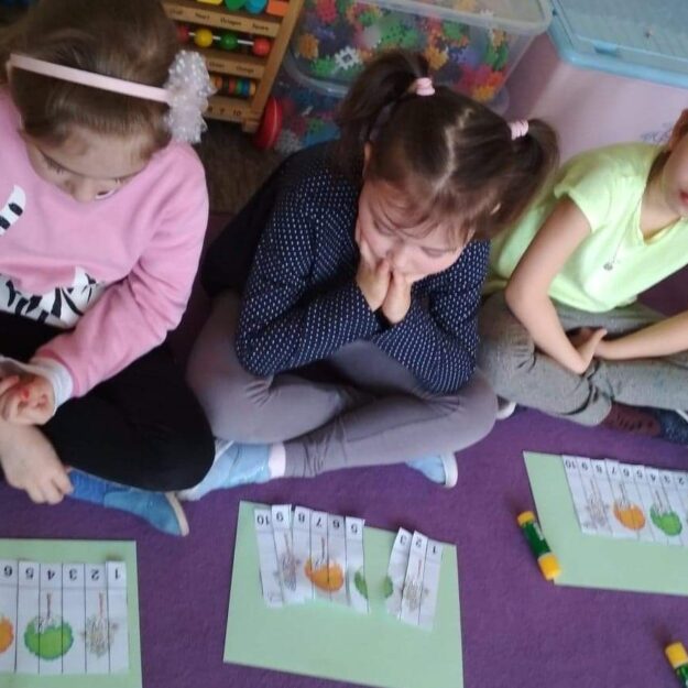 Dzieci siedzące po turecku na dywanie rozwiązują zadanie - układają pocięte paski papieru