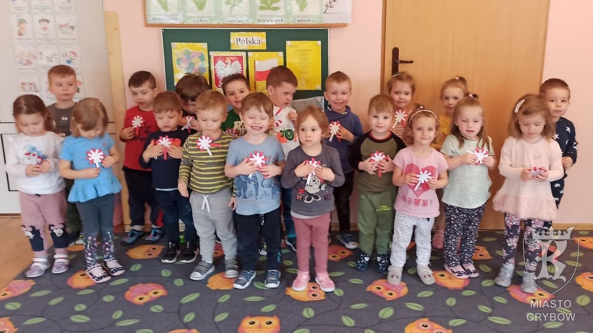 Dzieci stoją na dywanie w sali przedszkolnej, trzymają w rękach biało - czerwone kotyliony