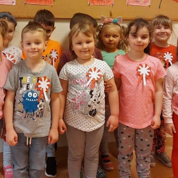 Dzieci stoją pod tablica z pracami plastycznymi i wskazują na wykonane przez siebie flagi Polski oraz mapy Polski