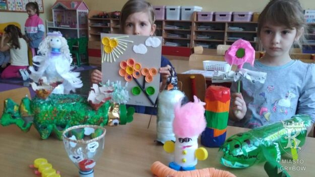 Na stoliku leżą prace plastyczne wykonane z odpadów, za nimi siedzą dwie dziewczynki i pokazują prace z plastikowych zakrętek