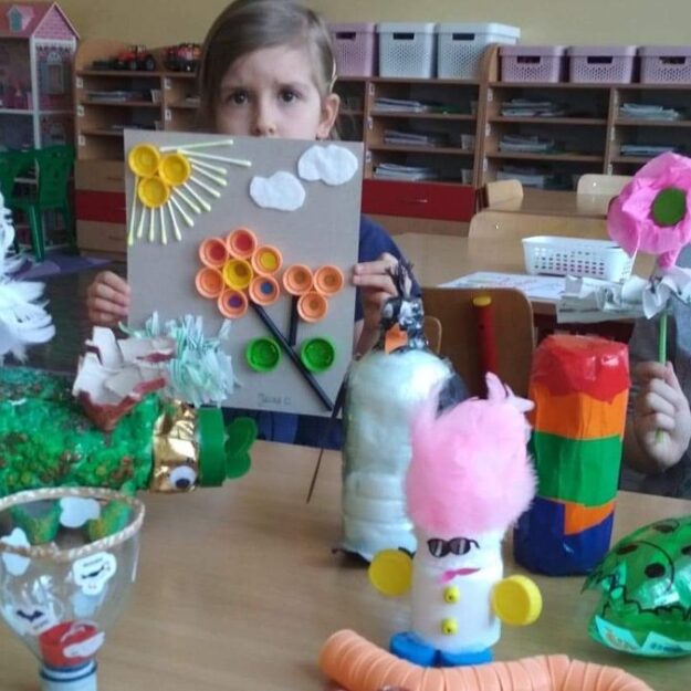 Na stoliku leżą prace plastyczne wykonane z odpadów, za nimi siedzą dwie dziewczynki i pokazują prace z plastikowych zakrętek