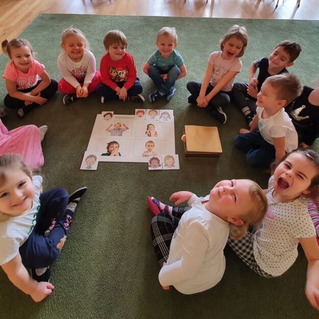 Dzieci siedzą w kole na dywanie - robią radosne miny. W środku koła karty demonstracyjne z emocjami i złote pudełko