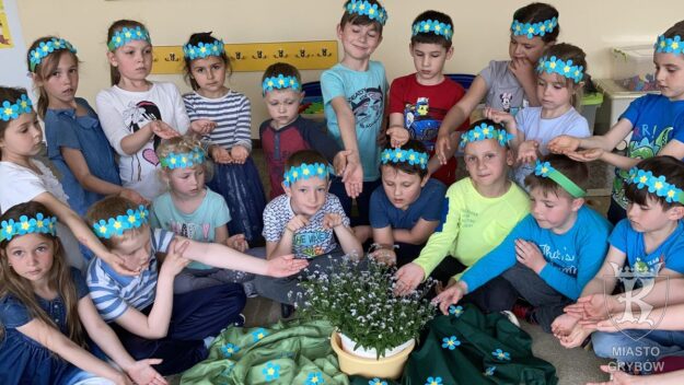 Ubrane na niebiesko dzieci siedzą wokół sadzonki kwiatków niezapominajek, na głowach opaski z kwiatkami niezapominajkami