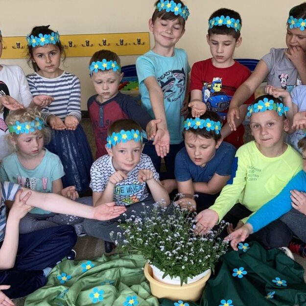 Ubrane na niebiesko dzieci siedzą wokół sadzonki kwiatków niezapominajek, na głowach opaski z kwiatkami niezapominajkami