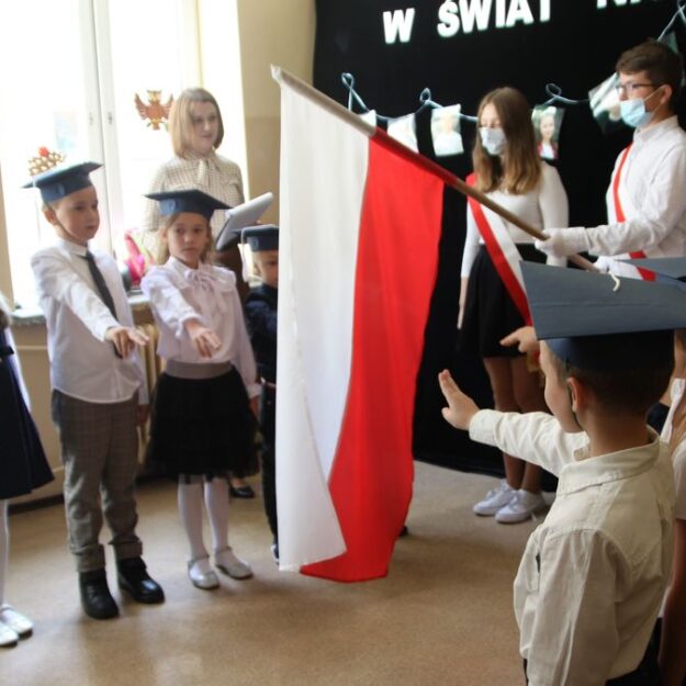 Moment ślubowania - dzieci z wystawionymi palcami do ślubowania, ustawione w dwóch rzędach, na środku pochylona flaga państwowa