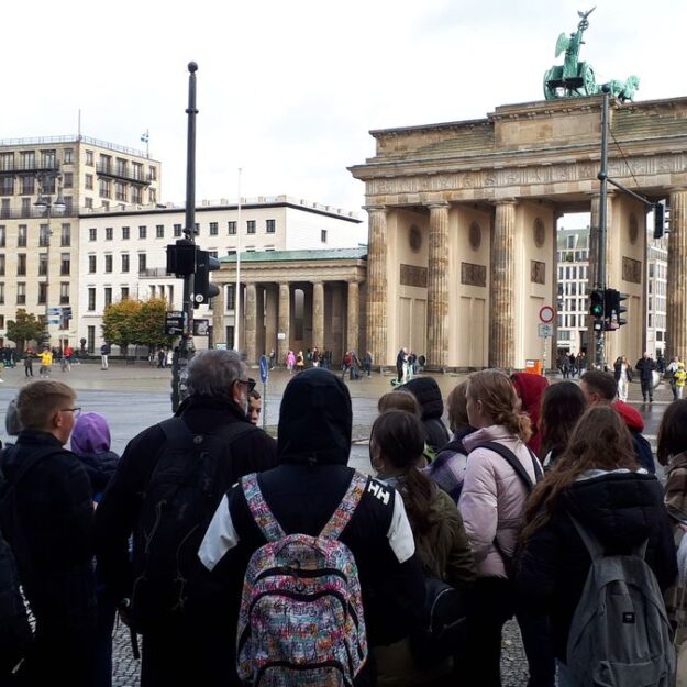 Na pierwszym planie grupa młodzieży słuchająca przewodnika, w tle widoczna Brama Brandenburska w centrum Berlina