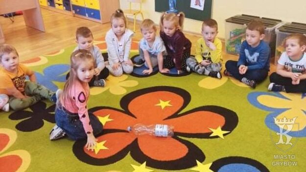Dzieci siedzą na dywanie w kole, na środku dziewczynka dotyka gwiazdkę z wróżbą andrzejkową, którą wylosowała