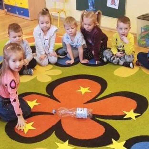 Dzieci siedzą na dywanie w kole, na środku dziewczynka dotyka gwiazdkę z wróżbą andrzejkową, którą wylosowała