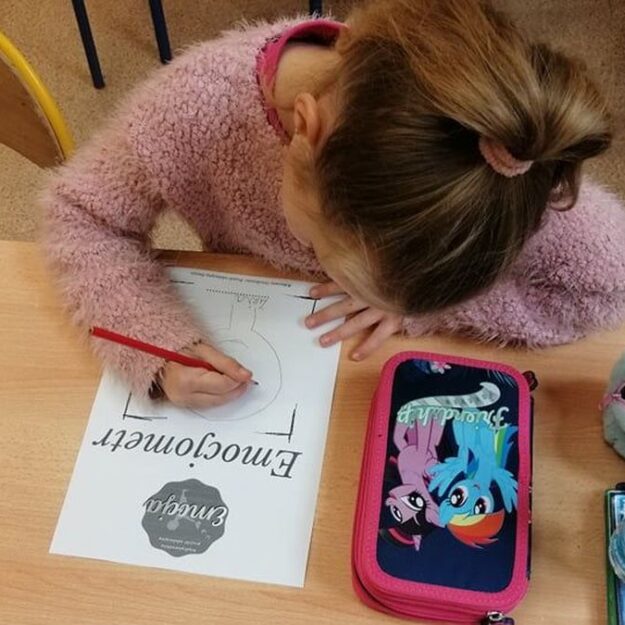 Dziewczynka wykonuje emocjometr - rysuje swój portret. Na ławce leżą jej książki i przybory szkolne