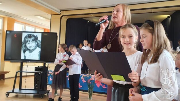Dzieci z nauczycielką trzymającą mikrofon, w tle ekran multimedialny