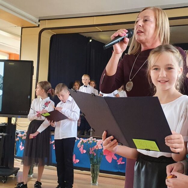 Dzieci z nauczycielką trzymającą mikrofon, w tle ekran multimedialny