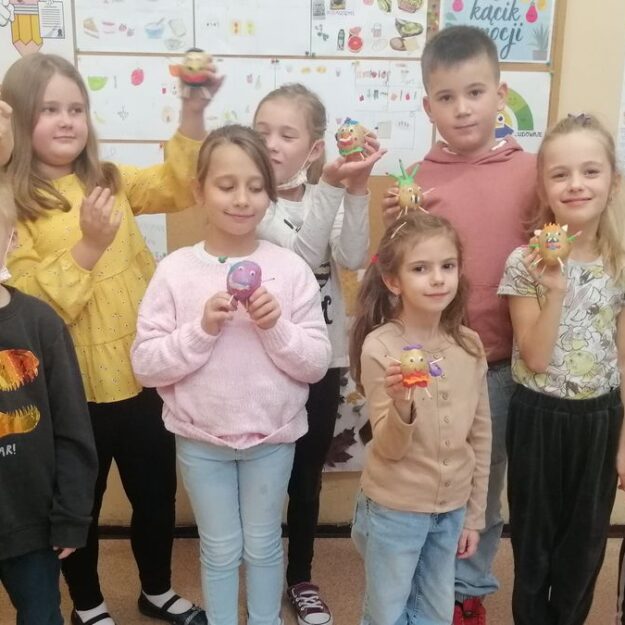 Dzieci stoją i trzymają w rękach wykonane z ziemniaków, zapałek i plasteliny cudaczki. W tle gazetka ścienna