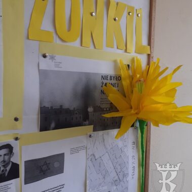 Tablica korkowa, na której umieszczone są informacje i zdjęcia historyczne. Z boku tablicy przypięty duży, żółty kwiat
