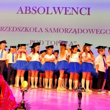 Dzieci stoją na scenie z biretami na głowie, z tyłu napis absolwenci