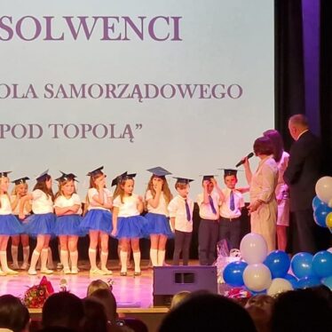 Dzieci stoją na scenie z biretami na głowie, z tyłu napis absolwenci