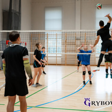 Mecz siatkówki - z lewej strony w wysokim wyskoku chłopak uderza w piłkę, reszta zawodników obserwuje jego akcję