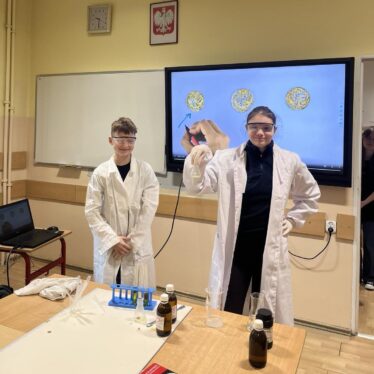 Dwójka uczniów chemików dumnie prezentuje zabarwiona fiolkę - z tyłu na monitorze interaktywnym trzy monety