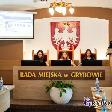 Widok w kierunku stołu prezydialnego, za którym siedzą przedstawiciele Młodzieżowej Rady, z prawej przemawiający Burmistrz, z lewej ekran z fragmentem prezentacji