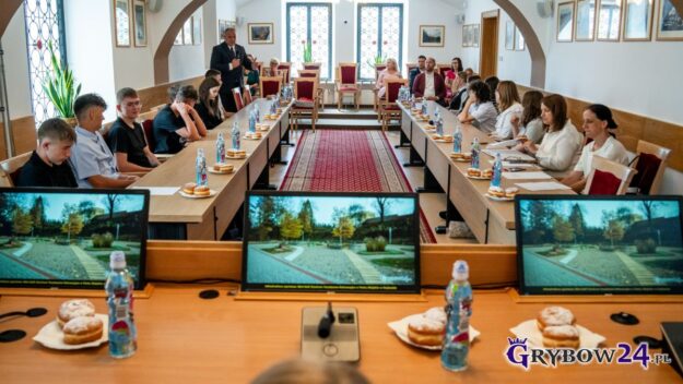 Widok na salę od stołu prezydialnego, na pierwszym planie trzy monitory komputerowe, w tle po obu stronach siedzi młodzież uczestnicząca w obradach. Z lewej przemawiający Burmistrz