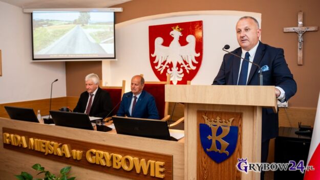 Burmistrz Miasta podczas swojej prezentacji, z lewej widok na ekran, pod nim przy stole prezydialnym siedzą Przewodniczący i Zastępca
