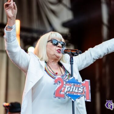 Główna solistka zespołu śpiewa do mikrofonu, na którym powieszony jest napis 2 plus 1