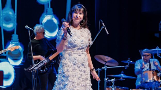 Uśmiechnięta Pani Krystyna Giżowska, trzymająca w prawej ręce mikrofon. W tle członkowie zespołu towarzyszącego