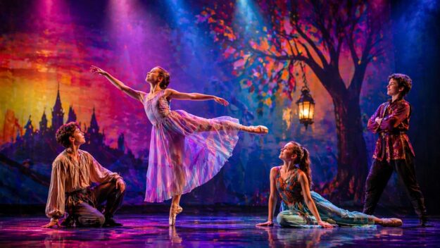 Kolorowa scena: czworo młodych tancerzy, baletnica w centrum w pozie arabeski, teatralne światła i bajkowe tło