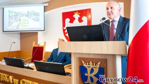 Burmistrz podczas podczas swojego sprawozdania przemawia do mikrofonu. W tle ekran z prezentacją